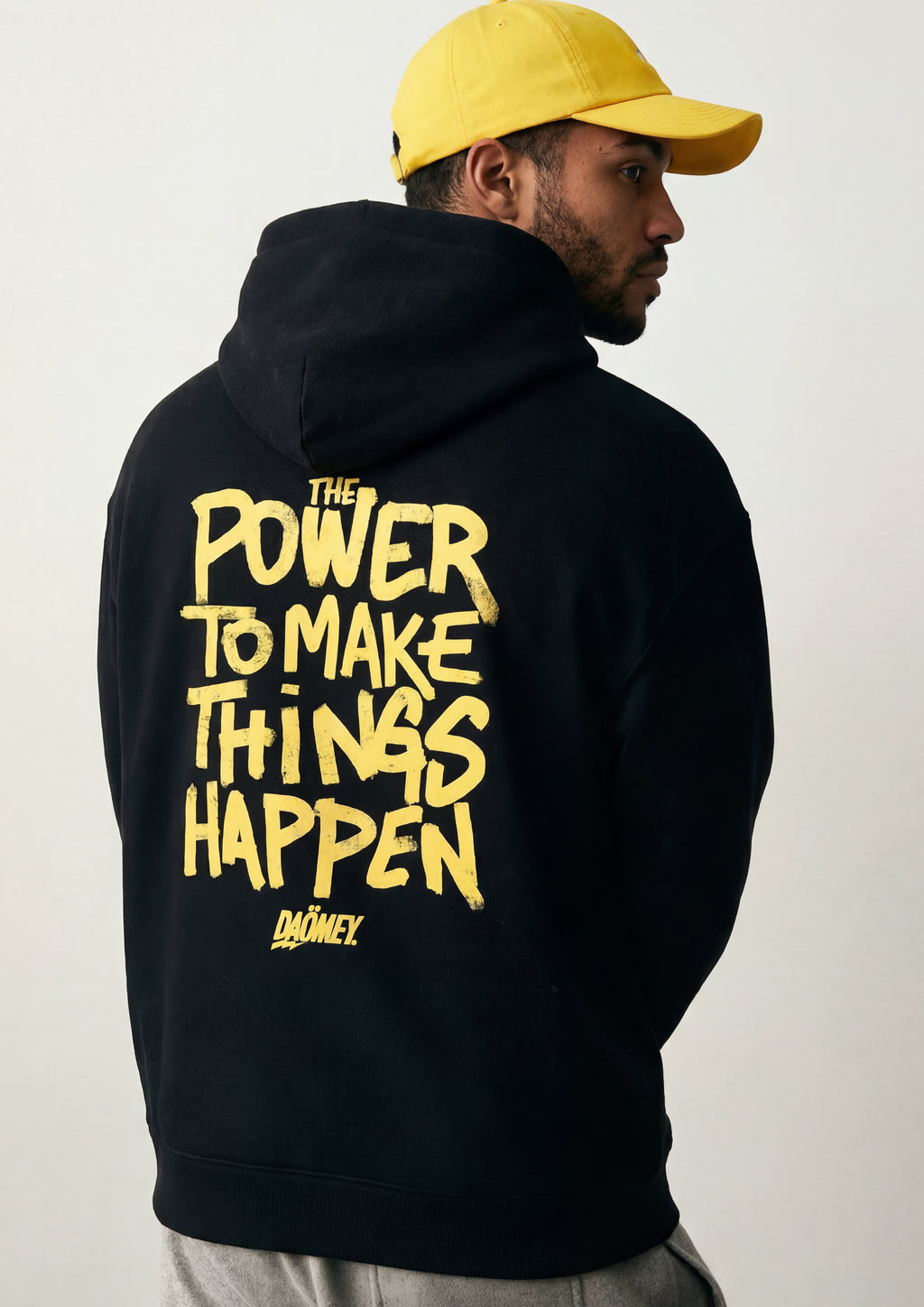 Person wearing a black hoodie with yellow text and a yellow cap on a white background